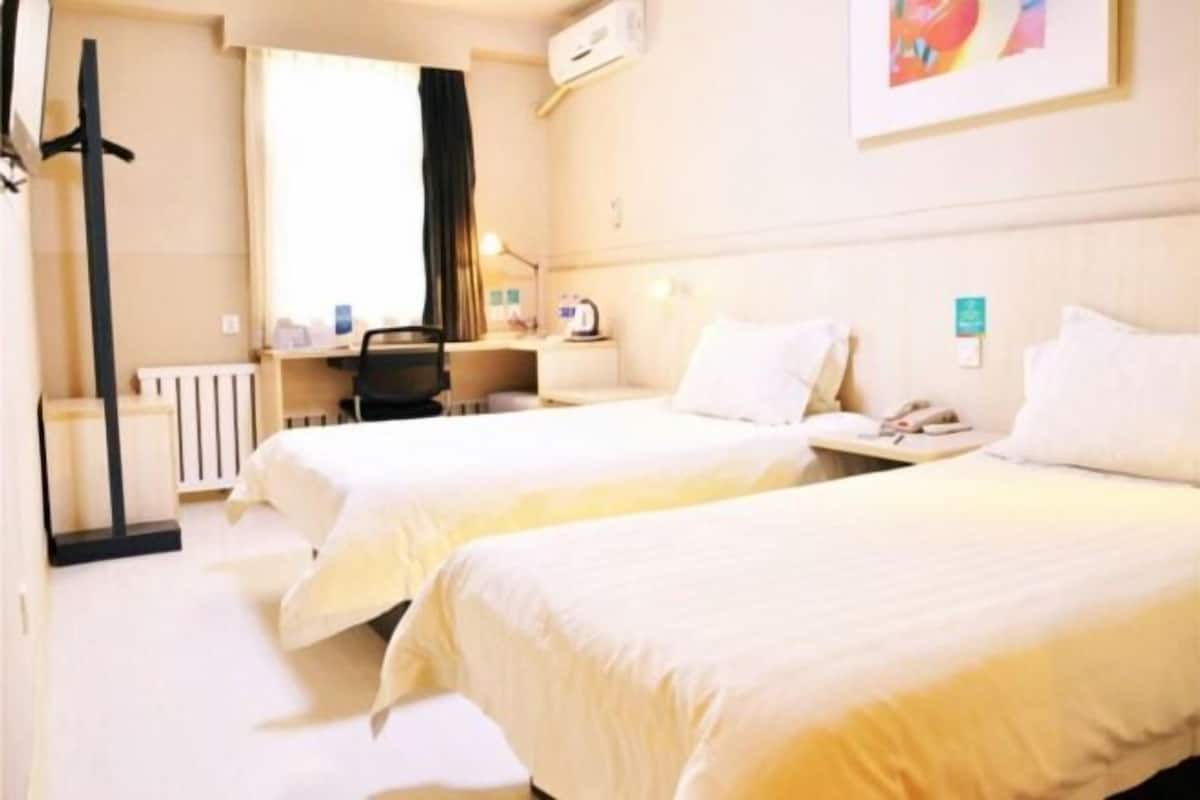 A hotel room with two beds, a desk, a chair, a radiator, and a window with curtains.
