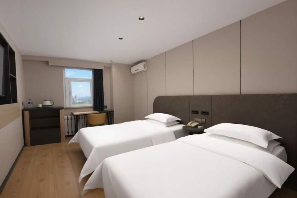 A hotel room with two beds, a desk, a chair, a window, and an air conditioning unit.