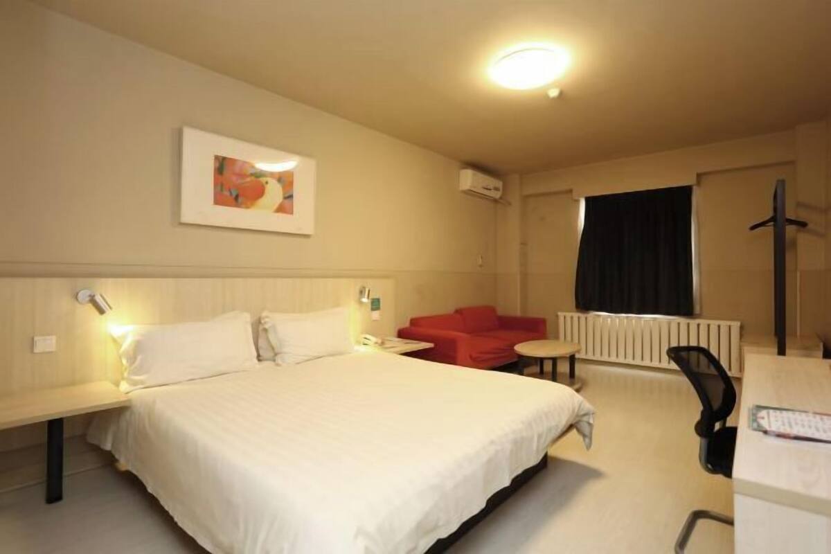 A hotel room with a bed, a red sofa, a small table, a desk, and a radiator.