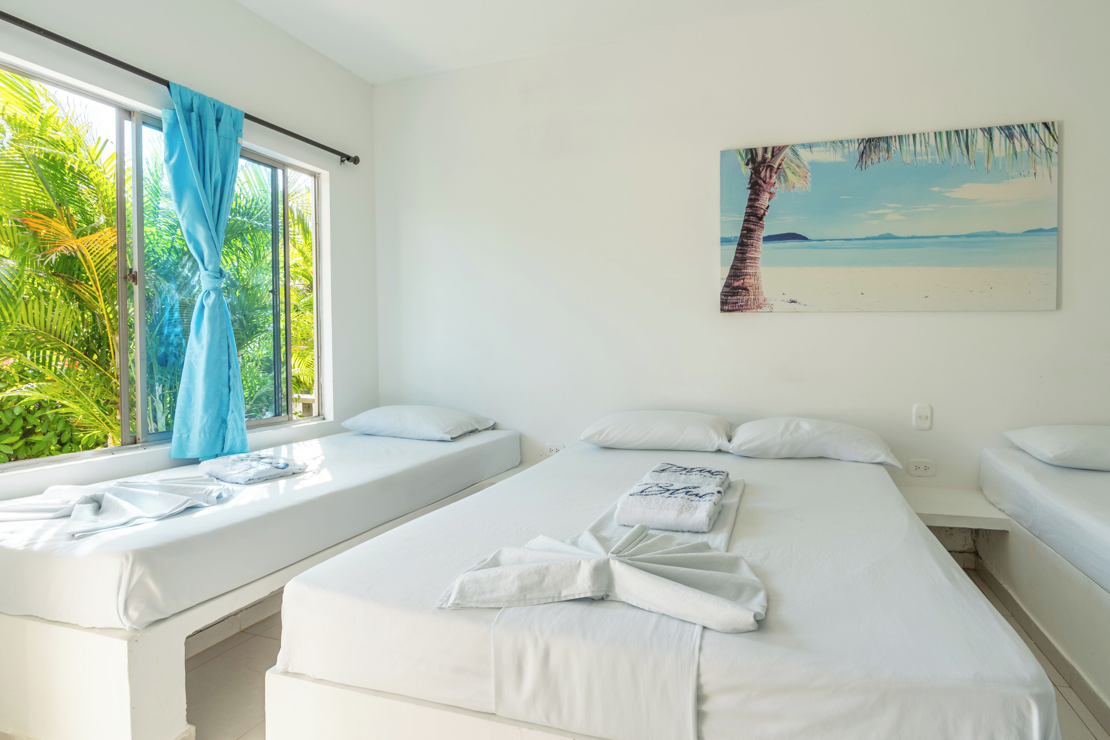 A hotel room with two beds, a window with curtains, a painting of a beach, and a view of greenery outside.