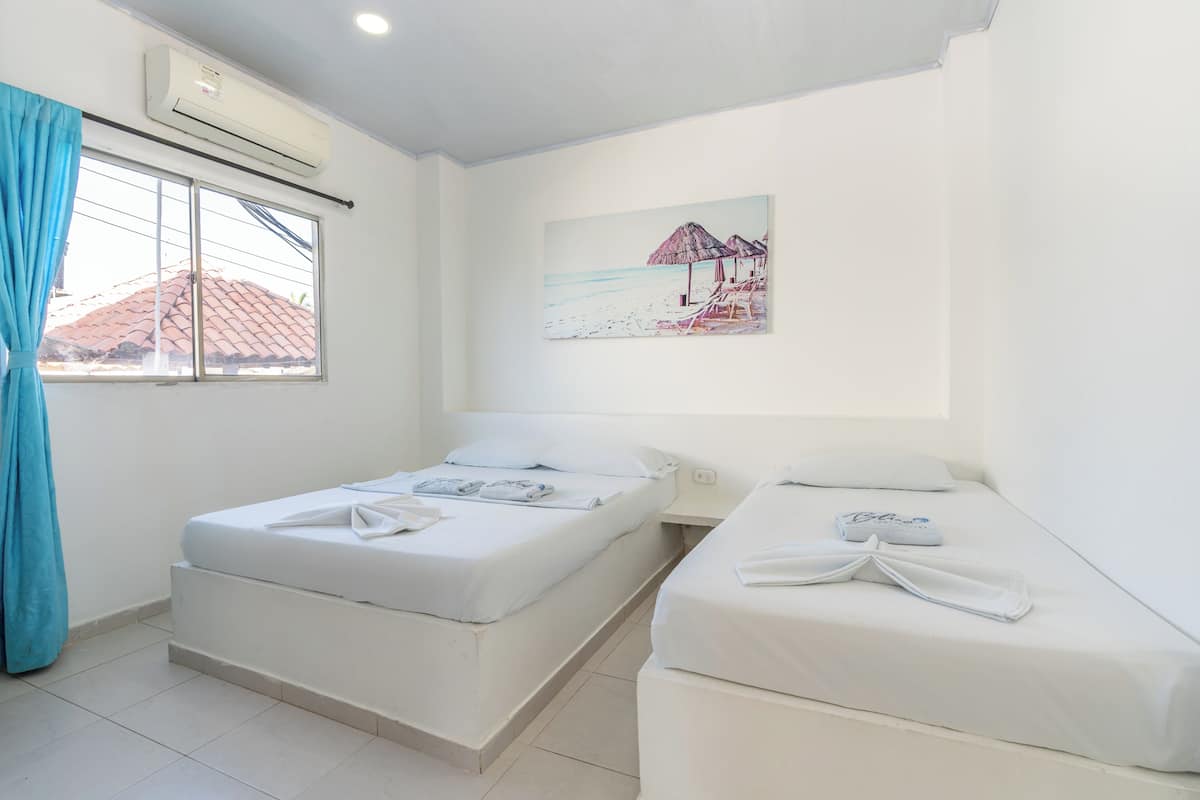 Una habitación con dos camas, una ventana con cortinas, aire acondicionado y un cuadro con motivos playeros.
