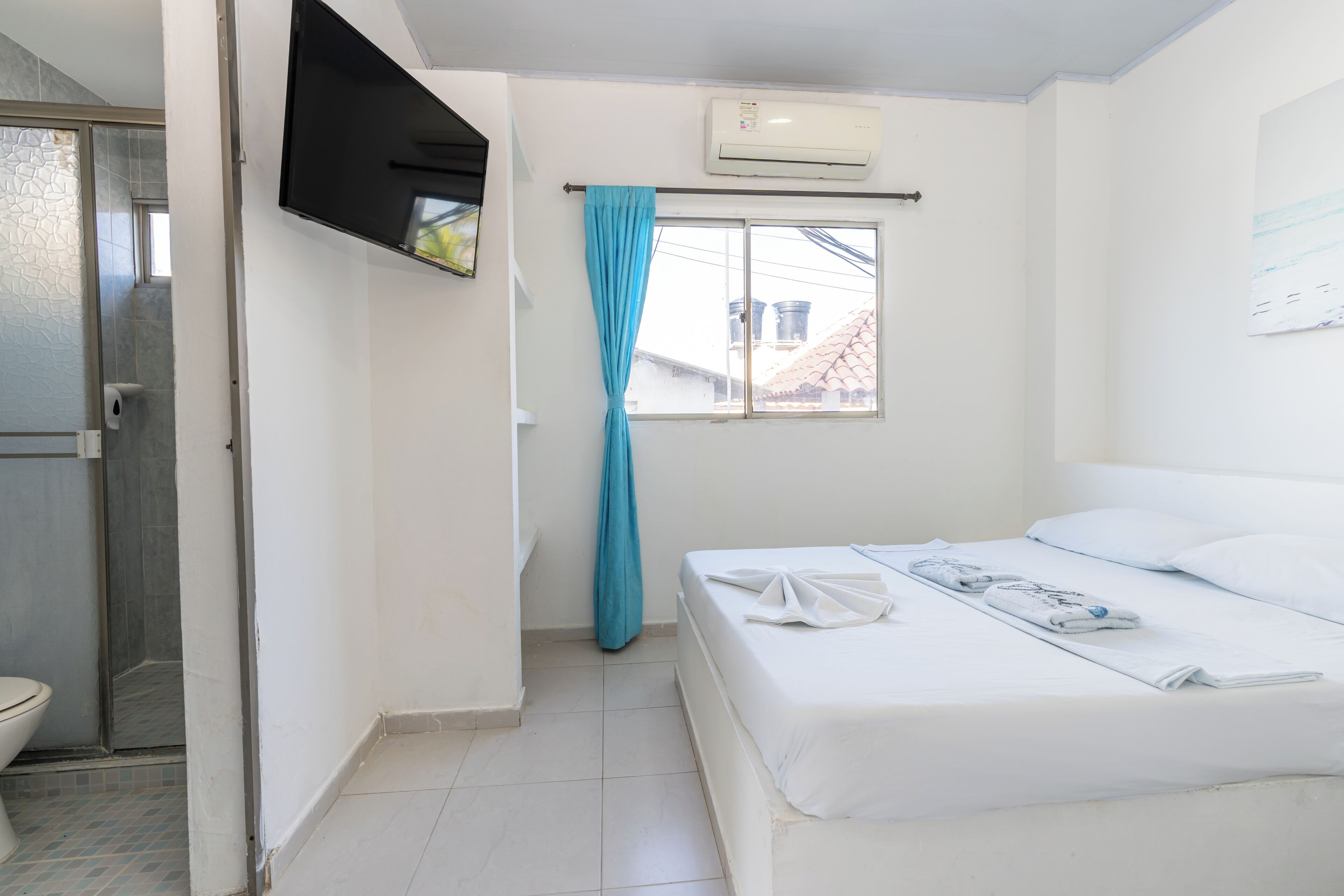 A hotel room with a bed, a TV mounted on the wall, a window with a curtain, and an air conditioning unit.