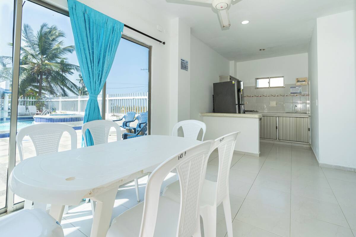 Habitación con mesa y sillas de comedor, una zona de cocina y vista a una piscina con palmeras.