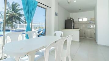 A room with a dining table and chairs, a kitchen area, and a view of a pool with palm trees.
