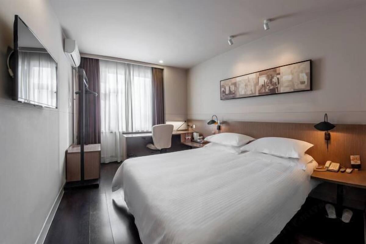 A hotel room with a bed, a desk, a chair, a mirror, a painting, and a window with curtains.
