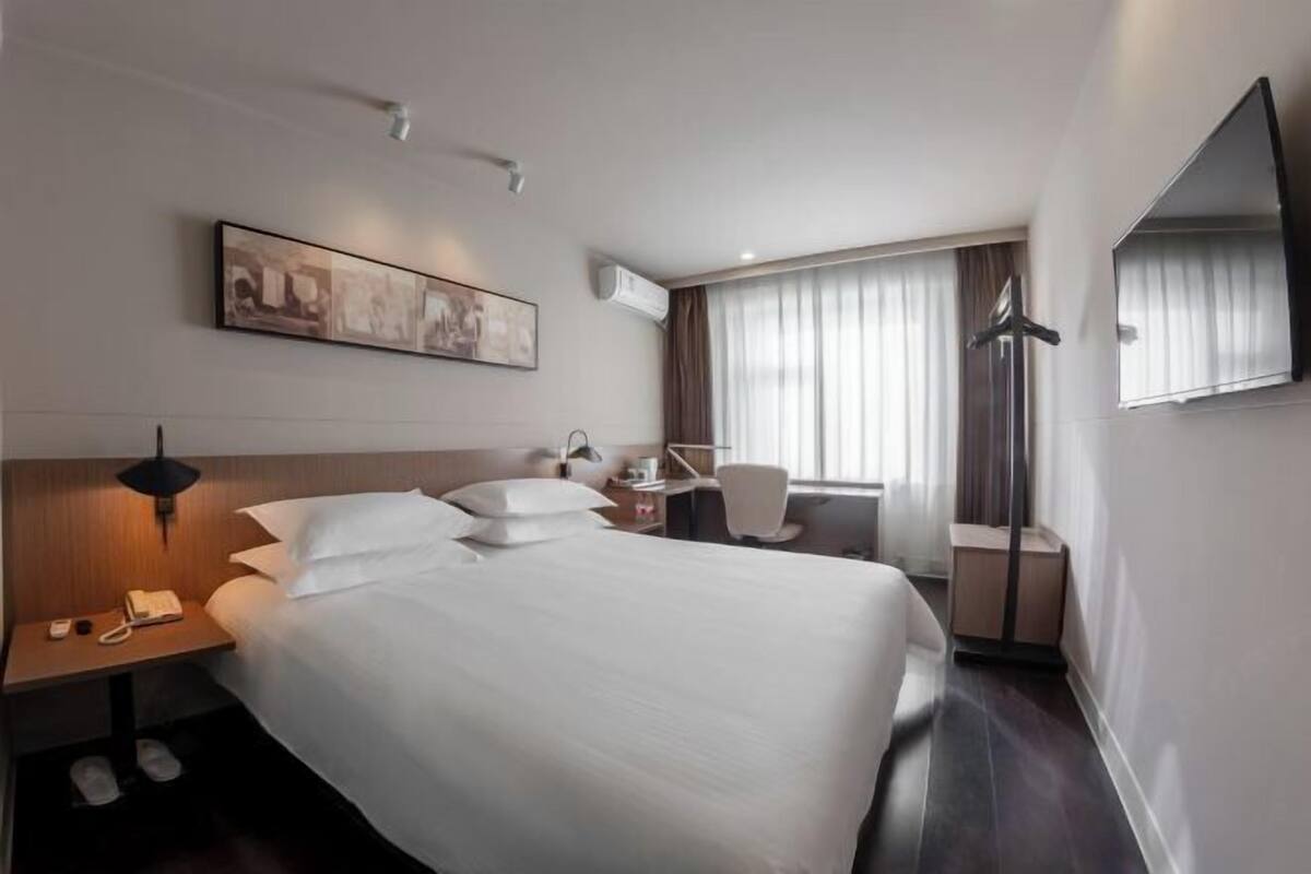 A hotel room with a large bed, bedside table with a lamp, a desk with a chair, a flat-screen TV, and a window with curtains.