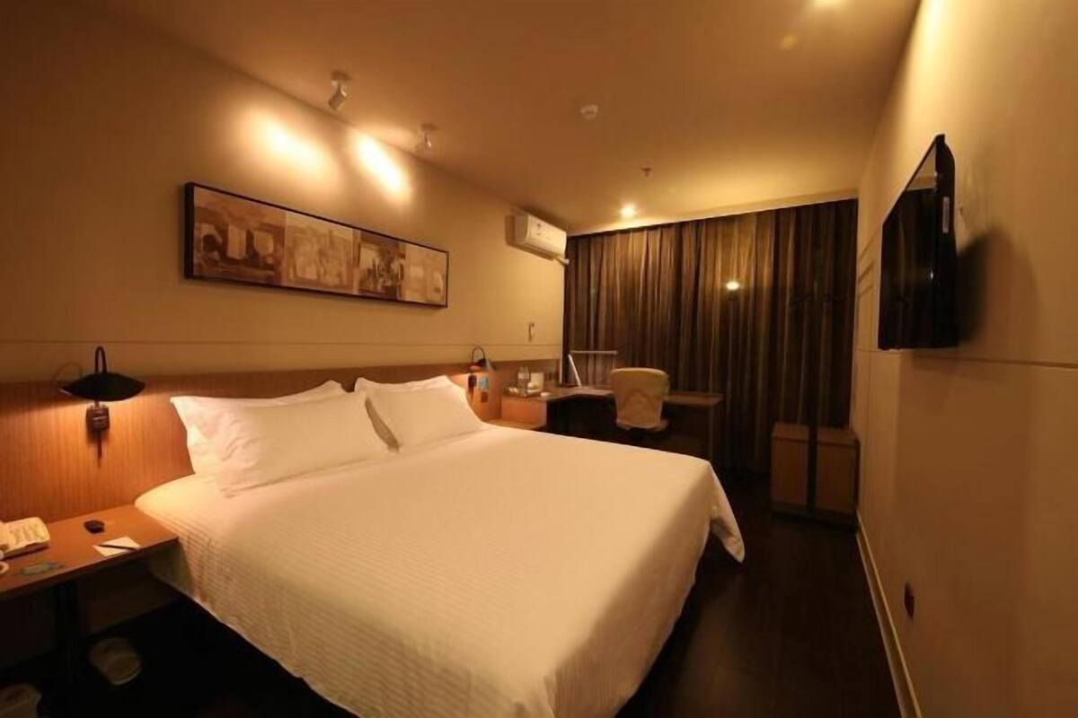 A hotel room with a large bed, a bedside table with a lamp, a chair, a desk, and a television mounted on the wall.