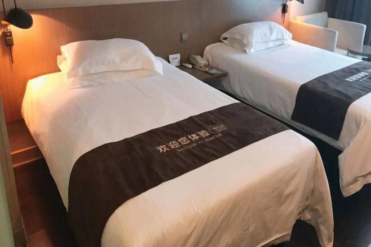 Two beds with white linens, a brown headboard, a nightstand with a phone, and a wall-mounted light.