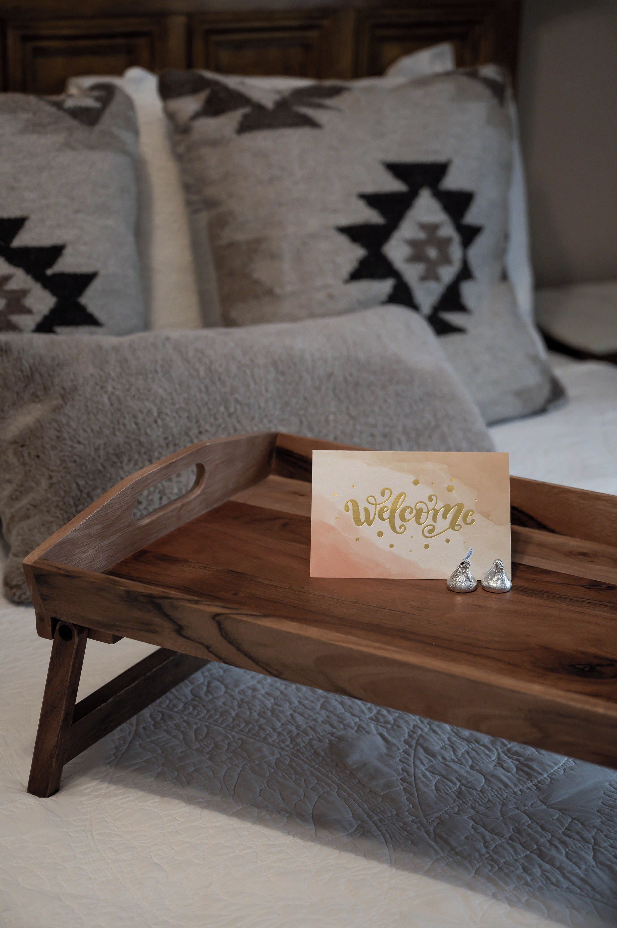 Ein hölzernes Tablett mit einer 'Welcome'-Karte auf einem Bett mit gemusterten Kissen.
