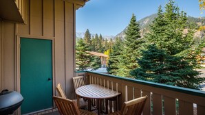 Uma varanda com mesa e cadeiras, uma porta verde, e vista para árvores e montanhas.