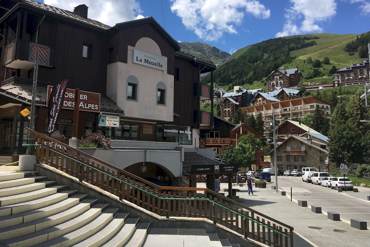 Ein Gebäude mit der Aufschrift 'La Muzelle', mit einer Treppe, die zu einer oberen Ebene führt, umgeben von weiteren Gebäuden und einer Bergkulisse.