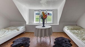 Habitació petita amb teulat inclinat, dos llits individuals, una taula i una finestra amb vista a un camp.
