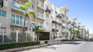 Un immeuble résidentiel à plusieurs étages, doté de balcons, d’une entrée sécurisée et de palmiers.