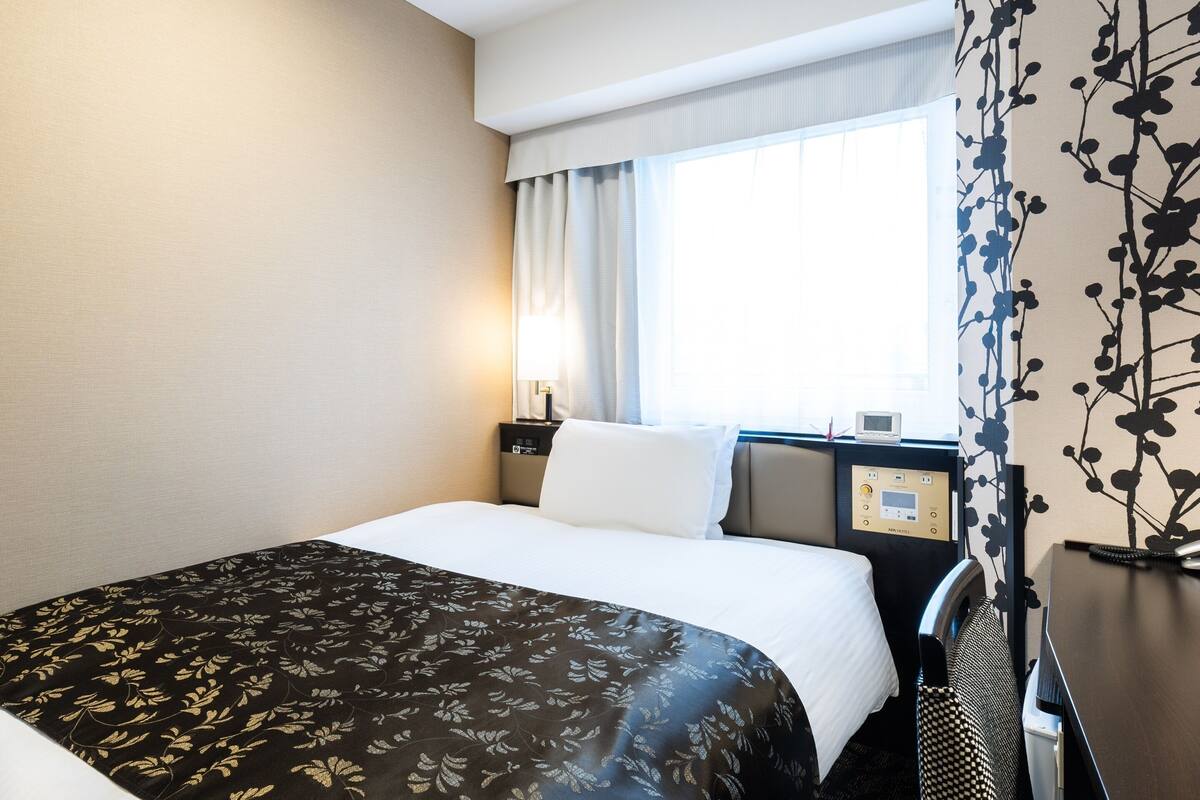 Une chambre d’hôtel avec un lit, une table de chevet avec une lampe, un panneau de commande et une fenêtre avec des rideaux.