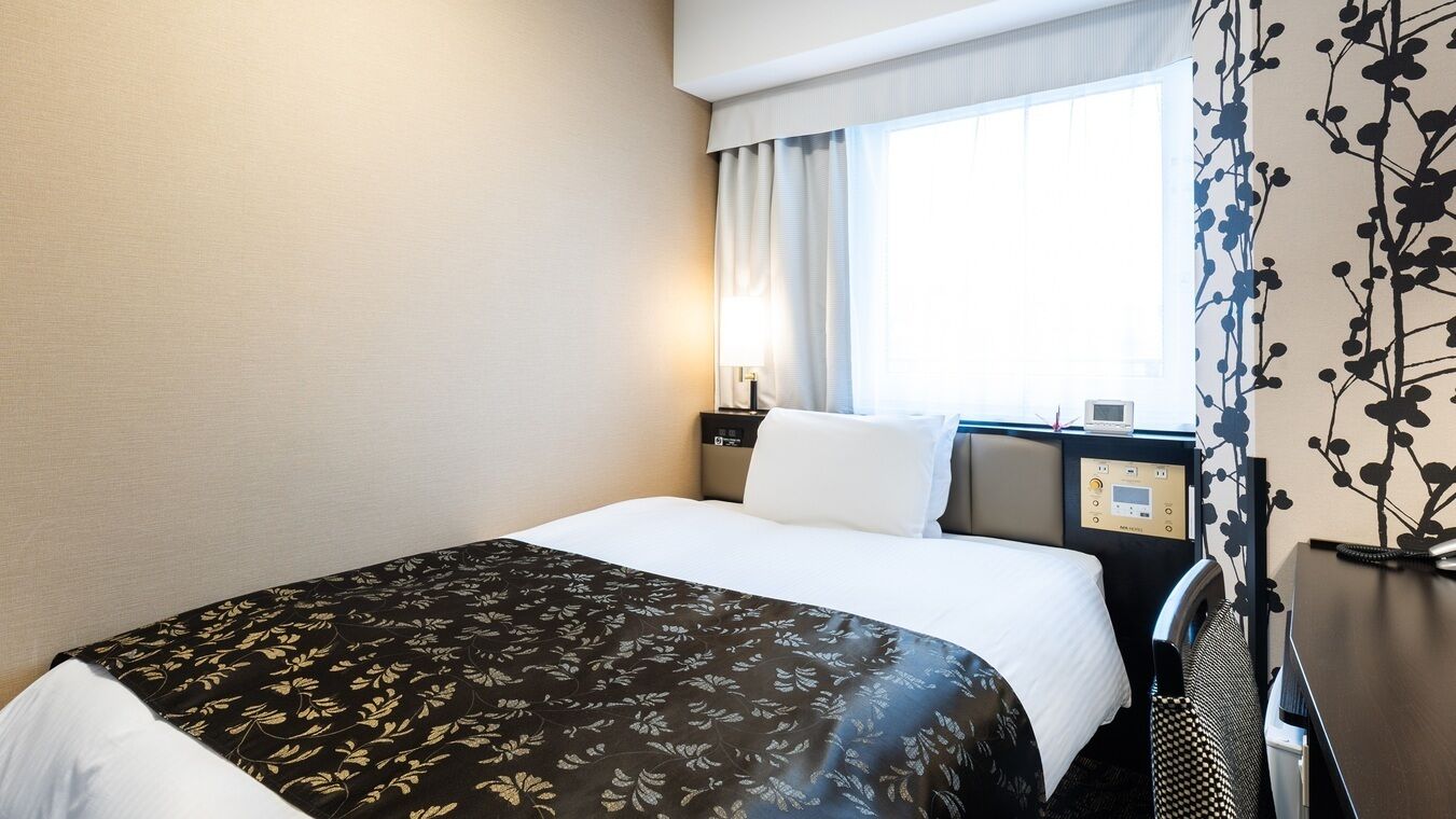 A hotel room with a bed, a bedside table with a lamp, a window with curtains, and a wall with a floral pattern.