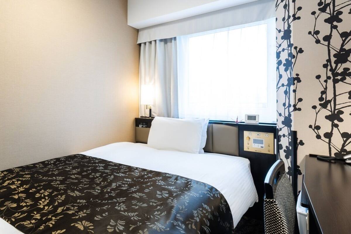 Une chambre d’hôtel avec un lit, une table de chevet avec une lampe, une fenêtre avec des rideaux et un mur orné d’un motif floral.