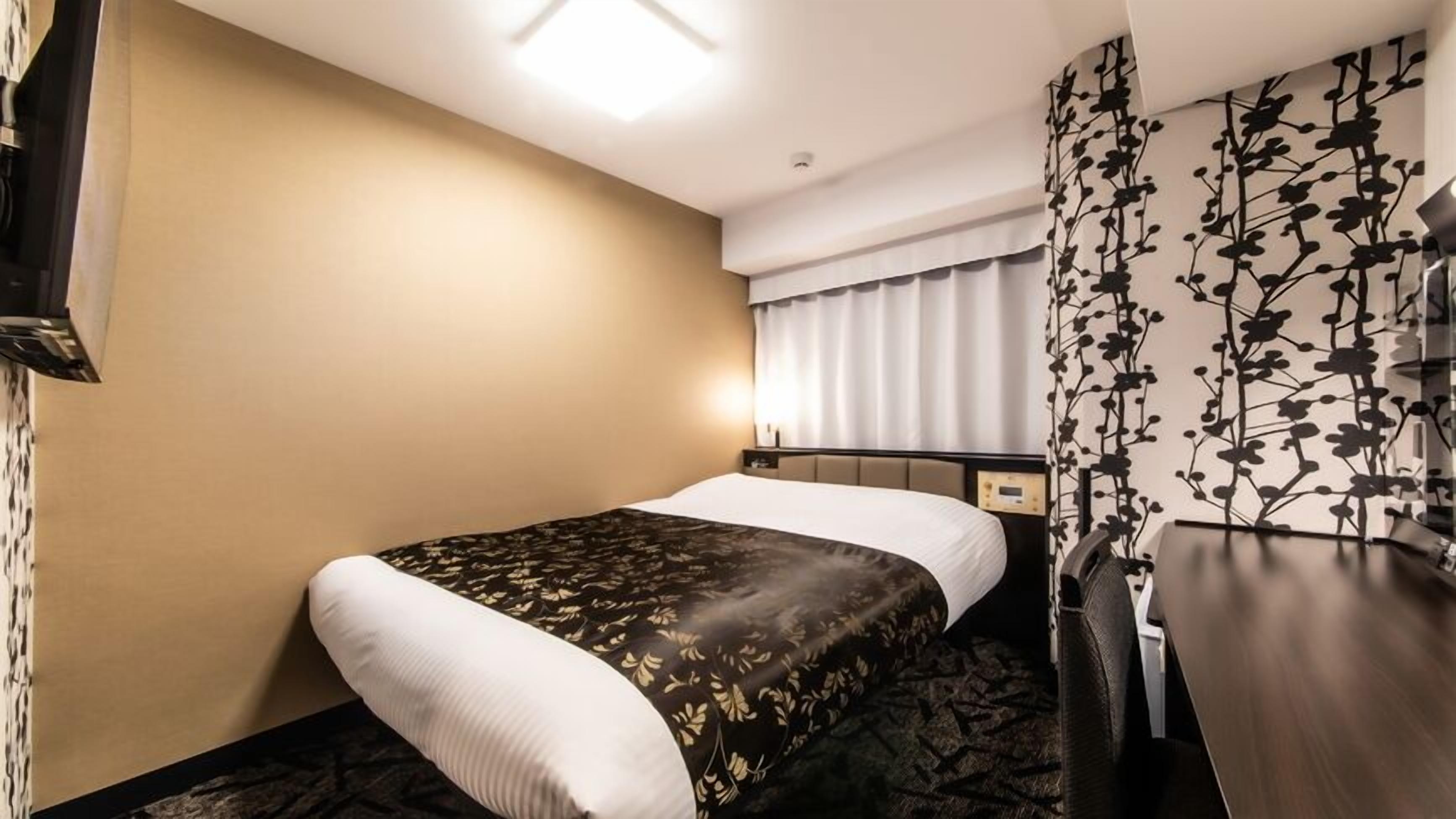A hotel room with a bed, a nightstand, a desk, a wall-mounted TV, and a window with curtains.