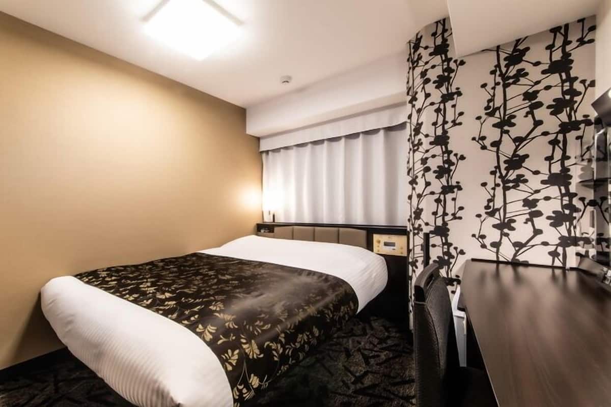 A hotel room with a bed, a nightstand, a desk, a wall-mounted TV, and a window with curtains.