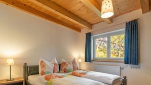 Ein Schlafzimmer mit Holzboden, einem Bett mit floralem Bettzeug, einem Nachttisch mit Lampe und einem Fenster mit Vorhängen.