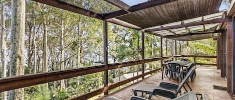 A covered outdoor seating area with tables and chairs, surrounded by a wooden deck and overlooking a forested area.