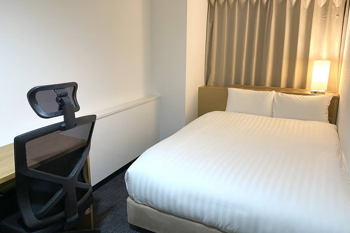 A hotel room with a bed, a desk with a chair, a nightstand, and a lamp.