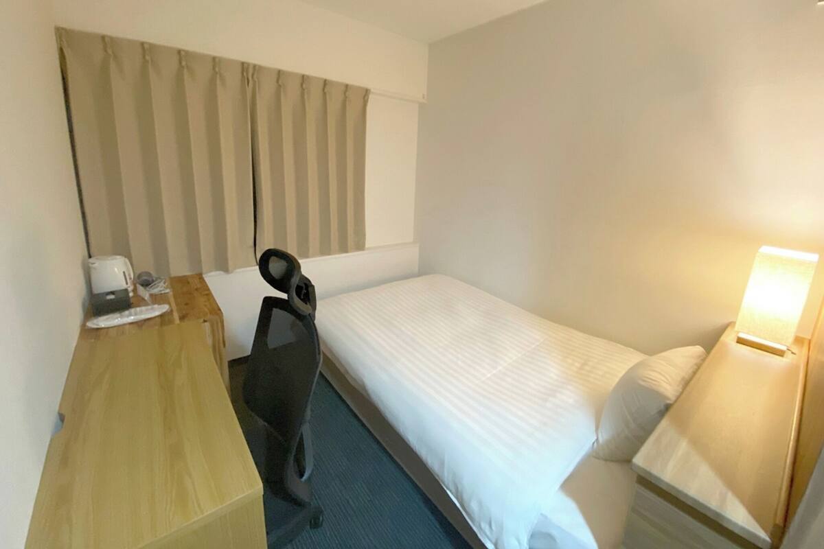 A hotel room with a single bed, a desk with a chair, a bedside table with a lamp, and a window with curtains.