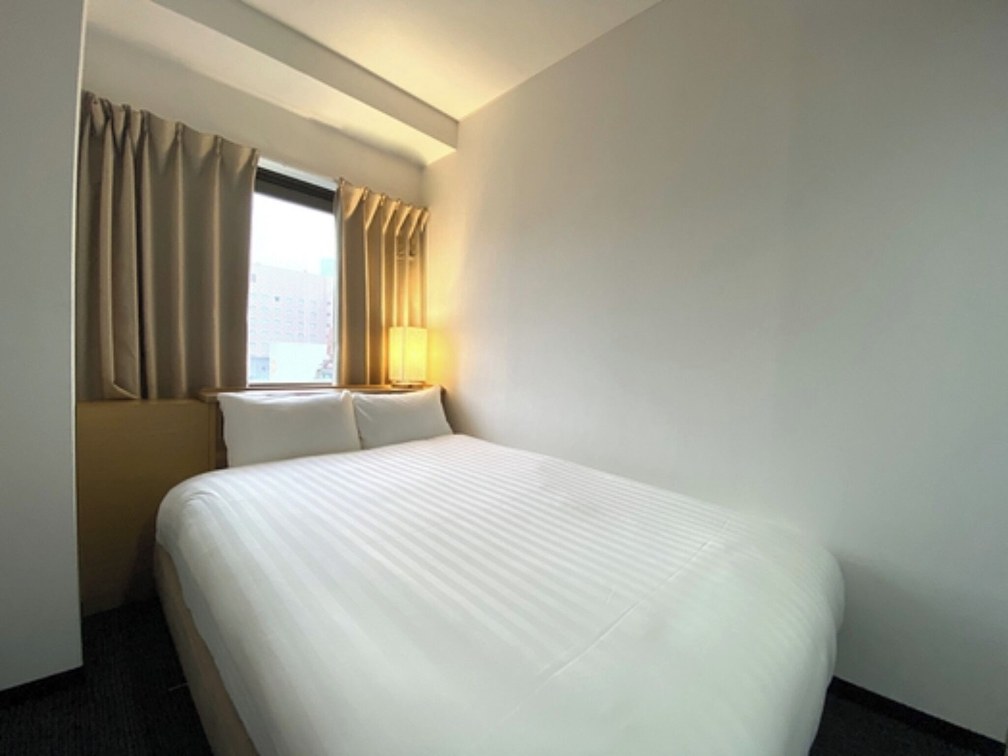 A hotel room with a large bed, a window with curtains, a nightstand with a lamp, and a view of the city.