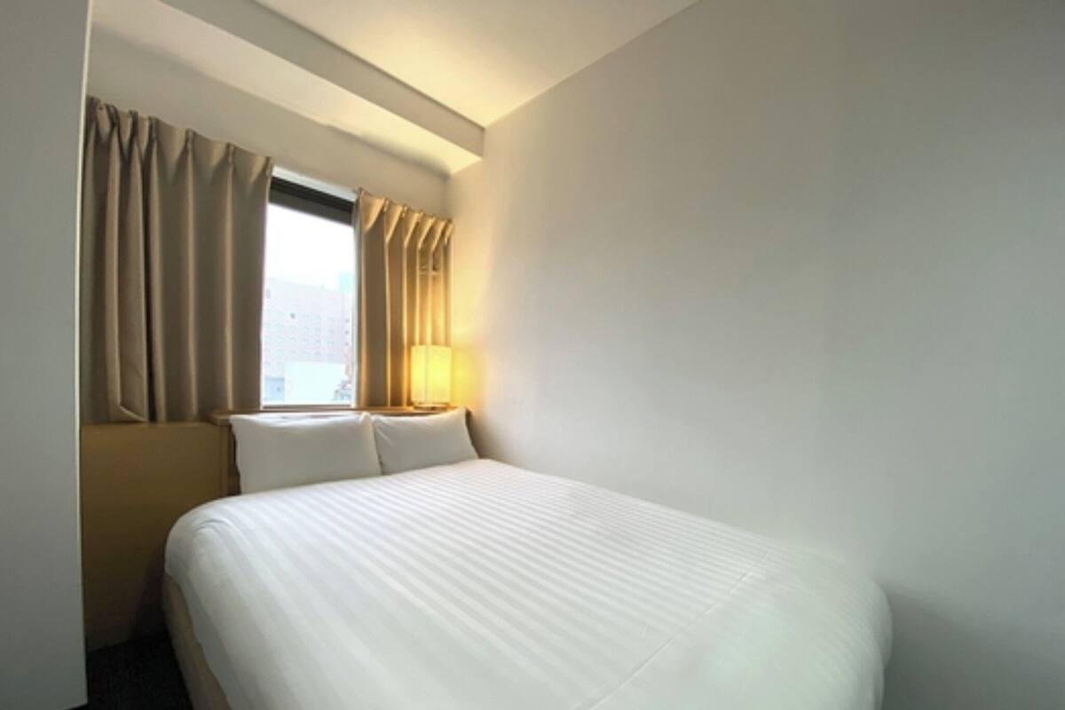 A hotel room with a large bed, a window with curtains, a bedside lamp, and a view of the city.