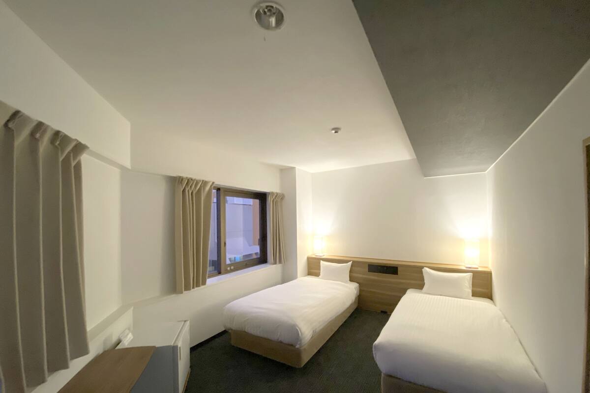 A hotel room with two single beds, a window with curtains, a desk, and a lamp.