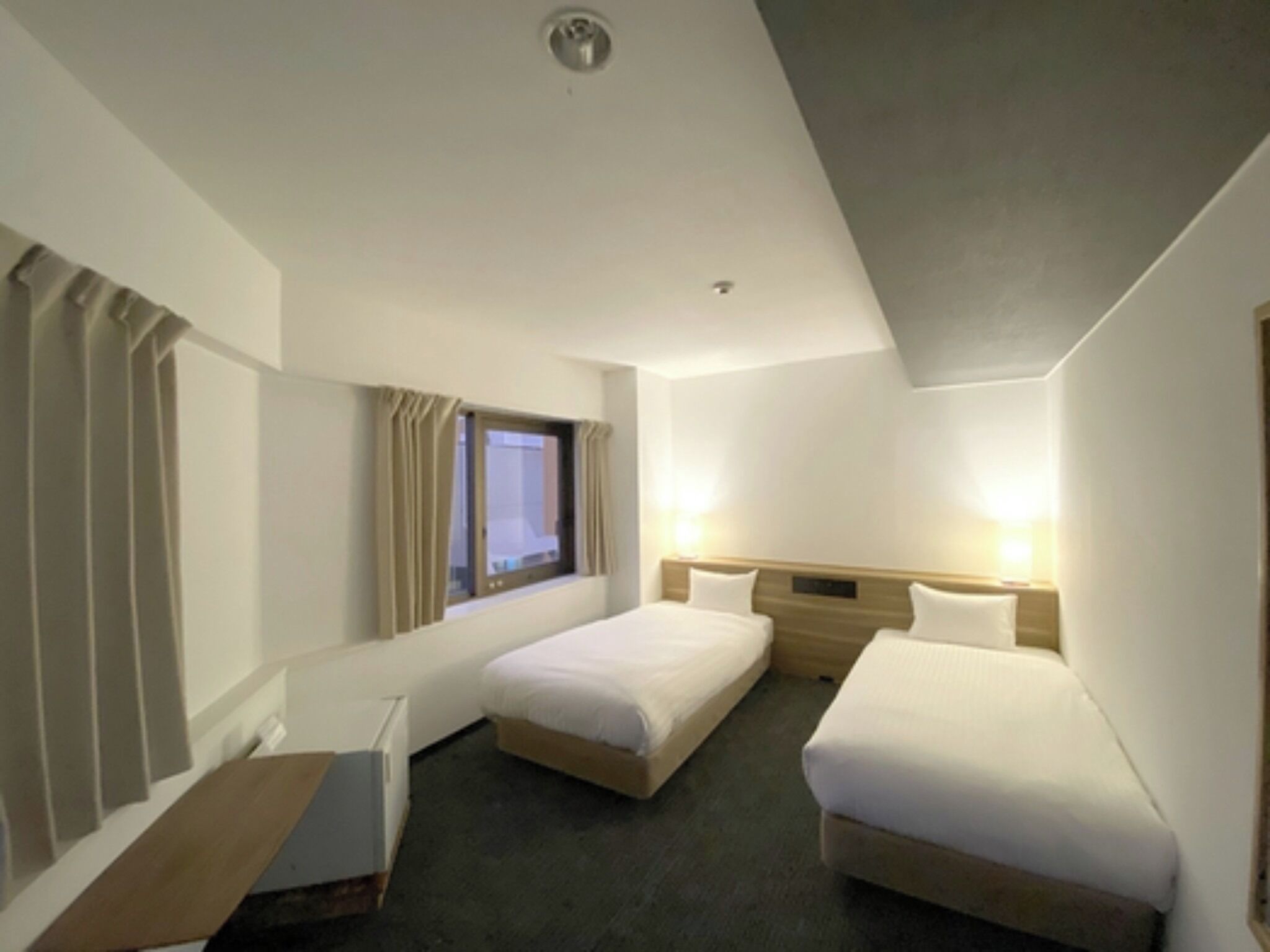A hotel room with two single beds, a window with curtains, a bedside table, and a wall-mounted light.