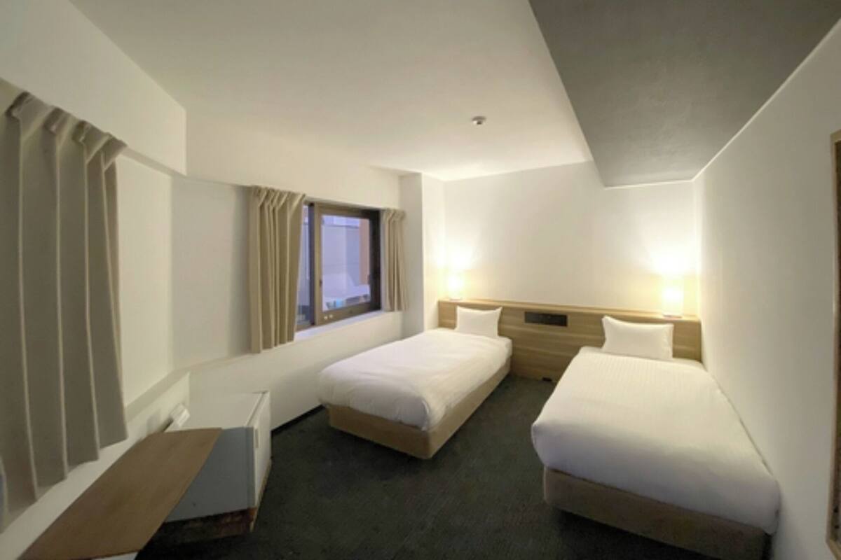 A hotel room with two single beds, a window with curtains, a desk, and a wall-mounted lamp.