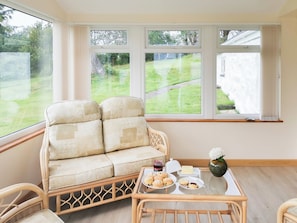 A room with a cushioned sofa, a small table with pastries, and large windows offering a view of a grassy area.