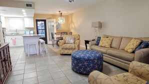 A living room with a sofa, ottoman, and armchair, a television on a cabinet, and a kitchen area in the background.
