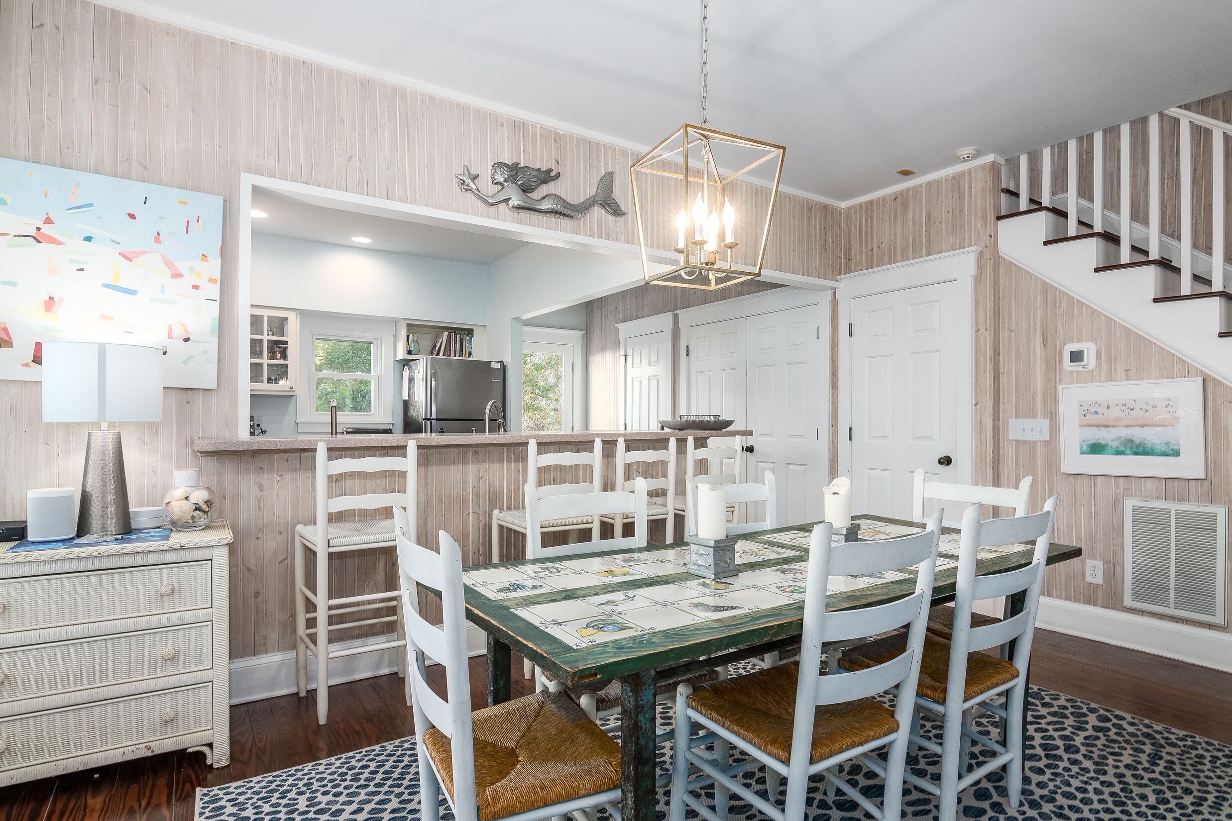 A dining area with a table set for six, surrounded by white chairs with woven seats. A chandelier hangs above the table.