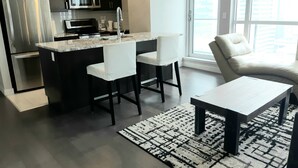 Eine moderne Küche mit dunklen Schränken, einem Edelstahlkühlschrank, einer Mikrowelle und einer Kaffeemaschine. Eine kleine Kücheninsel beherbergt zwei weiße Stühle und einen Tisch. Das Zimmer verfügt über ein großes Fenster mit Blick auf Gebäude.