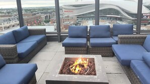Eine Dachterrasse mit blauen Sitzgelegenheiten, einem zentralen Feuerkorb und Blick auf ein modernes Stadion.