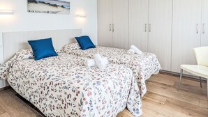 A bedroom with two beds, a wooden wardrobe, a painting on the wall, and a white chair.