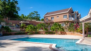 Una piscina con borde de baldosas, rodeada de palmeras y una casa grande de fachada marrón y tejado rojo.