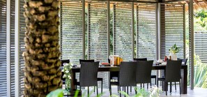 Un espace de restauration en plein air avec une table dressée pour quatre personnes, entourée de chaises en osier noir, et une grande fenêtre dotée de stores horizontaux.