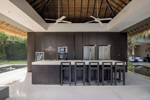 A modern kitchen with a central island, dark cabinetry, and a thatched ceiling. - Sangsuri Villa 2 (Koh Samui)