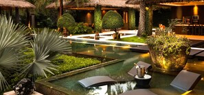 Un espace piscine extérieur luxueux, doté d’une structure à toit de chaume, d’un coin salon avec une bouteille de champagne et d’une grande plante en pot.