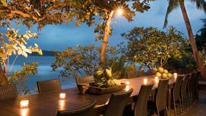 An outdoor dining area with a wooden table and chairs, surrounded by tropical plants and overlooking a body of water. - Sangsuri Villa 2 (Koh Samui)