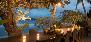 An outdoor dining area with a wooden table and chairs, surrounded by tropical plants and overlooking a body of water. - Sangsuri Villa 2 (Koh Samui)