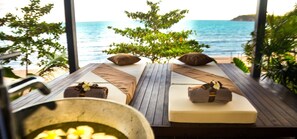 Un salón junto a la playa con suelo de madera, dos camas reclinables con almohadas y toallas, un cuenco grande de frutas y vistas al océano.