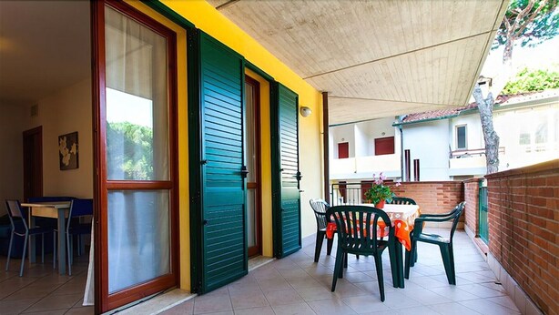 A covered patio with a table and chairs, a sliding door, and a view of the street. - Lido Spina / Estensi Apartments (Comacchio)