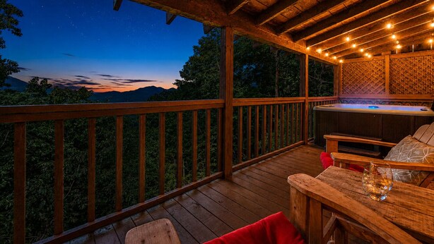 Une terrasse en bois avec un jacuzzi, des sièges et une vue sur les montagnes au crépuscule.