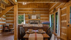L’intérieur d’un chalet en bois, avec une table à manger dressée pour deux personnes, une cheminée et une vue sur l’extérieur.