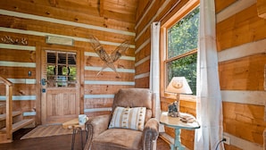 L’intérieur chaleureux d’un chalet, avec un décor en bois, un fauteuil moelleux agrémenté d’un coussin à motifs, une petite table avec une lampe et une fenêtre donnant sur un paysage verdoyant.