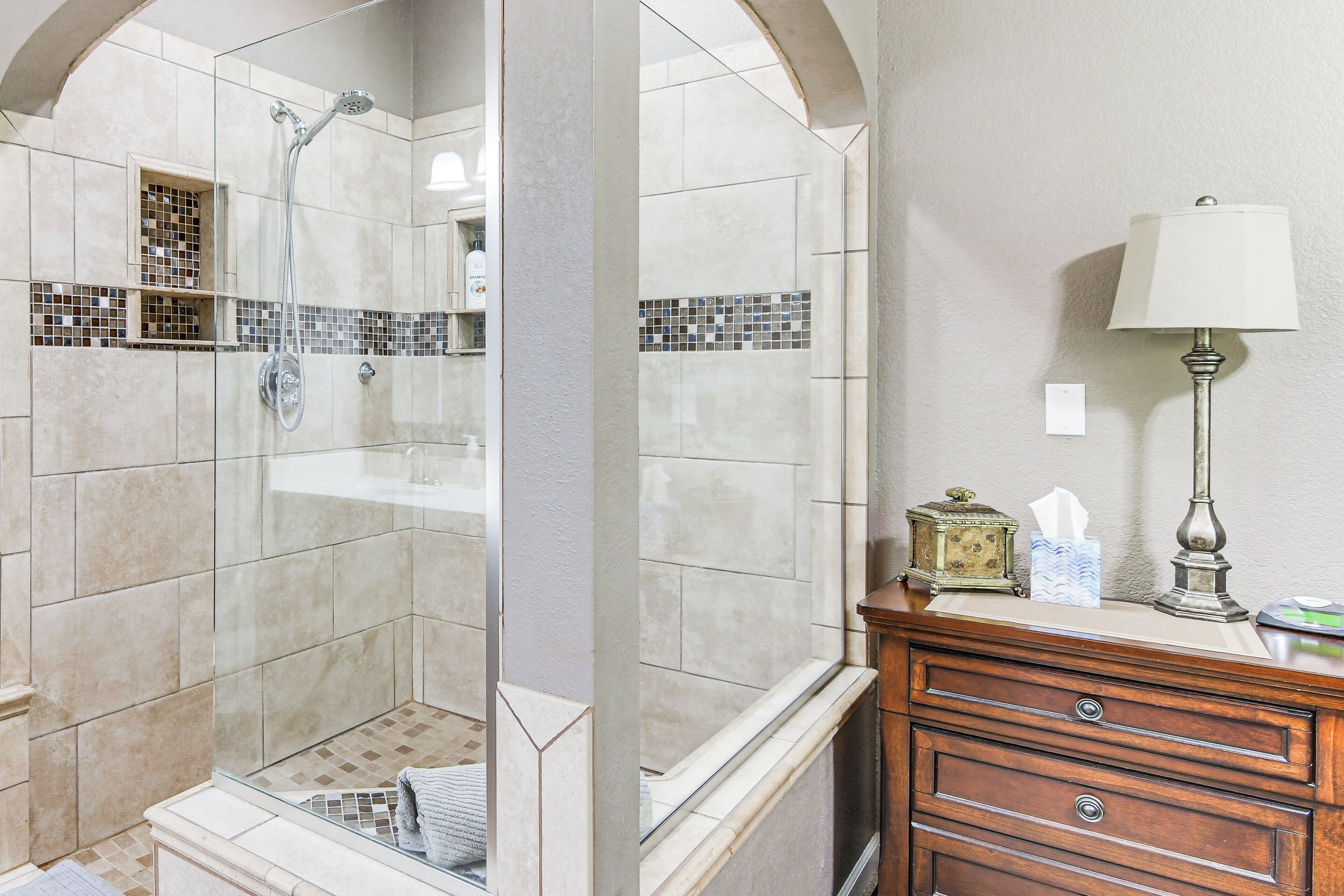 A bathroom with a glass-enclosed shower, a wooden vanity with a mirror, and a wall-mounted lamp.