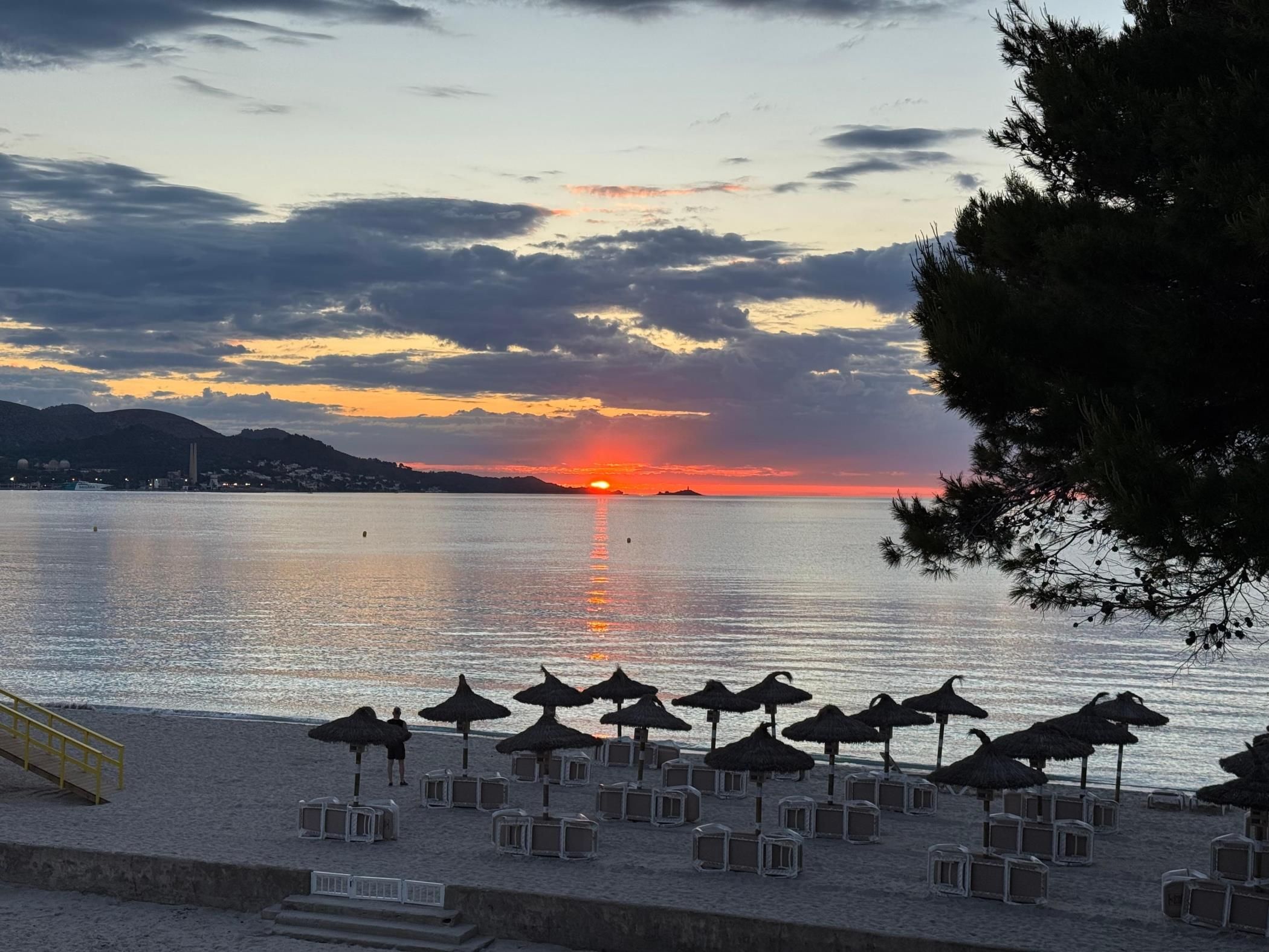 Una platja amb ombrel·les i gandules, un sol ponent sobre l'aigua i una ciutat al capdamunt.